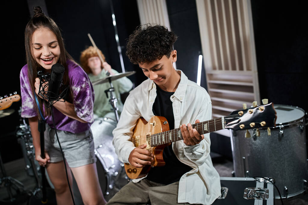 veselé roztomilé dospívající dívka zpívající vedle svého roztomilého přítele hrající na kytaru a další chlapec na bicí - Fotografie, Obrázek