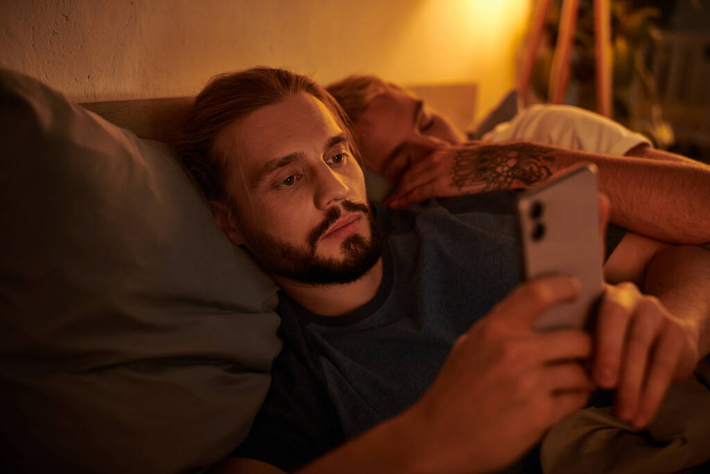 illoyaler bärtiger Schwuler surft Date-App auf Smartphone in der Nähe schlafenden Freundes in der Nacht im Schlafzimmer - Foto, Bild