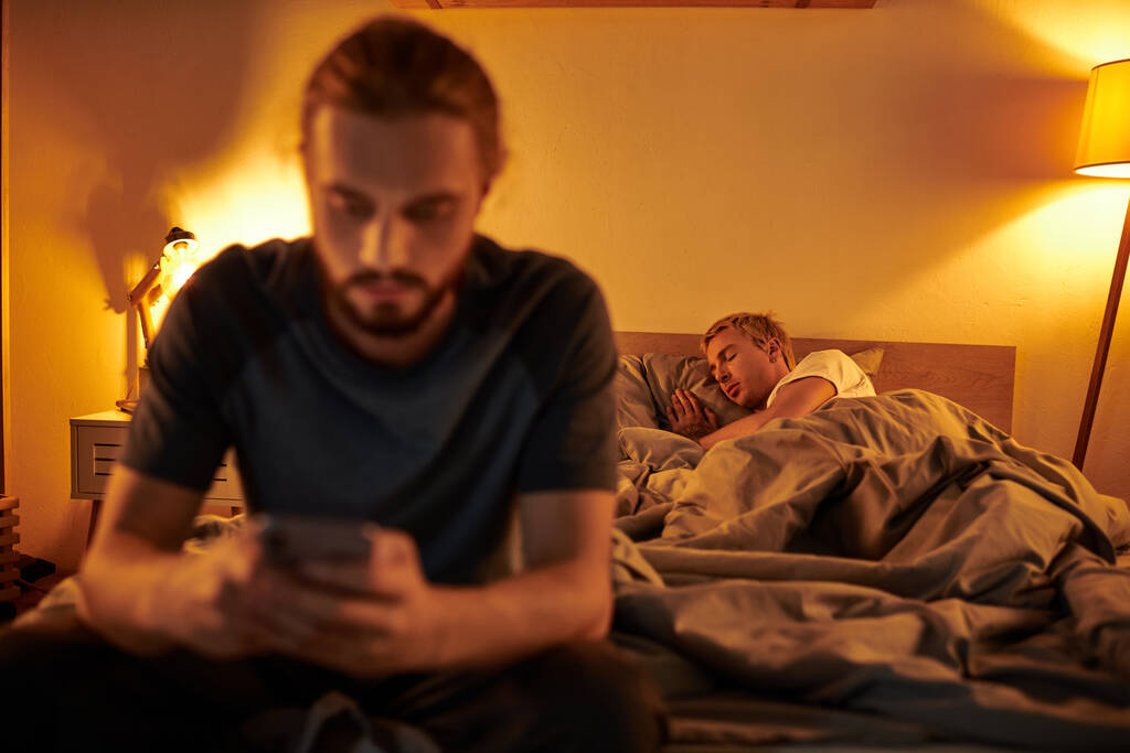 disloyal bearded gay man browsing internet on smartphone near partner sleeping at night in bedroom - Photo, Image