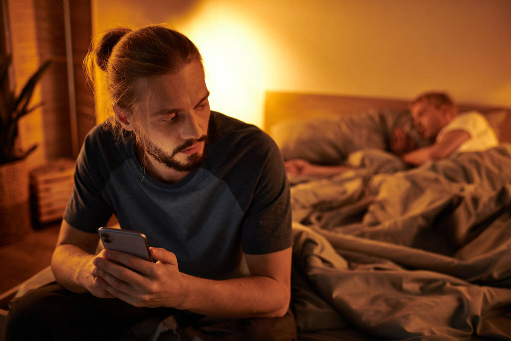 disloyal bearded gay man browsing date app on smartphone near partner sleeping at night in bedroom - Photo, Image