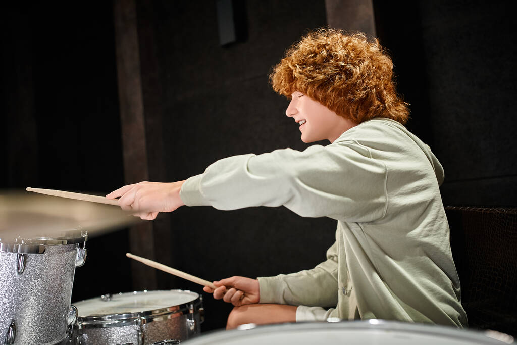 veselé rozkošné červené vlasy dospívající chlapec v ležérní útulný outfit hrát jeho bicí aktivně ve studiu - Fotografie, Obrázek