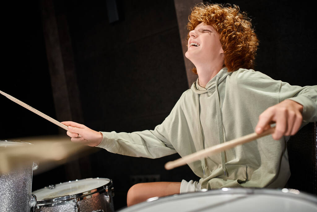 vreugdevolle getalenteerde rood harige tiener in comfortabele dagelijkse kleding spelen van zijn drums terwijl in de studio - Foto, afbeelding
