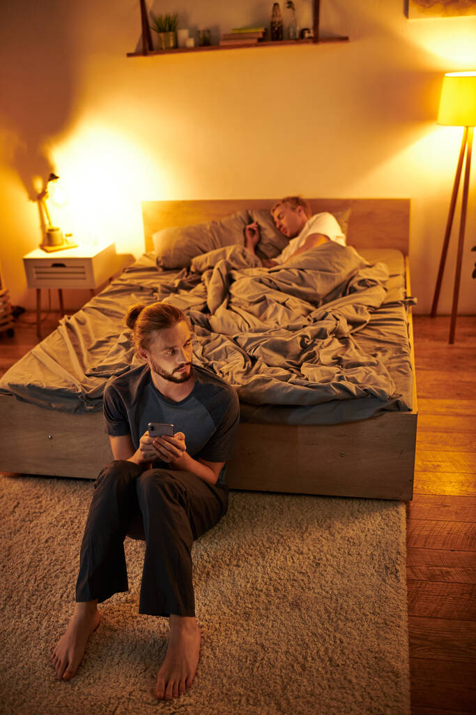 nielojalny brodaty gej człowiek przeglądanie data app w pobliżu śpiący chłopak w nocy w sypialnia, zdrada - Zdjęcie, obraz