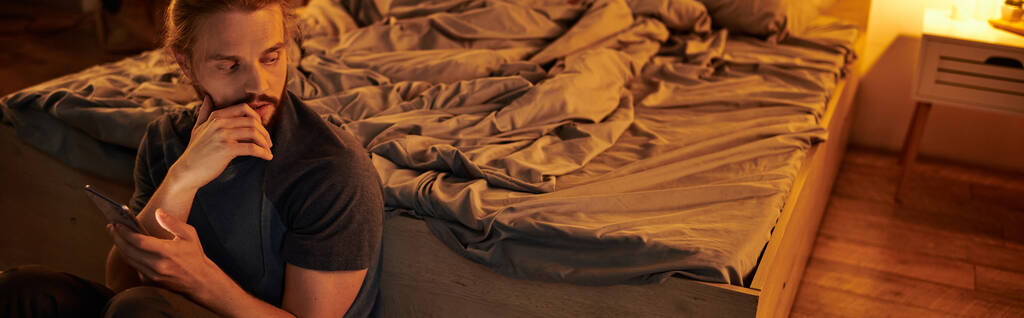 γενειοφόρος στοχαστικός άνθρωπος με κινητό τηλέφωνο κάθεται κοντά στο κρεβάτι στο υψηλό στην κρεβατοκάμαρα, οριζόντια banner - Φωτογραφία, εικόνα
