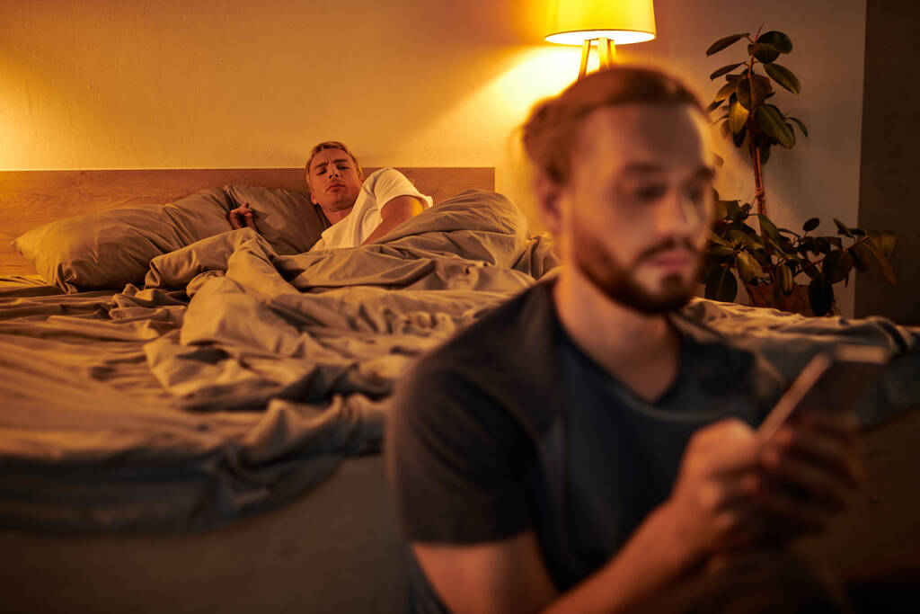 αποθαρρυμένος γκέι άντρας που κοιτάζει το αγόρι της να στέλνει μηνύματα στο κινητό τηλέφωνο στο υπνοδωμάτιο το βράδυ, εξαπατώντας - Φωτογραφία, εικόνα