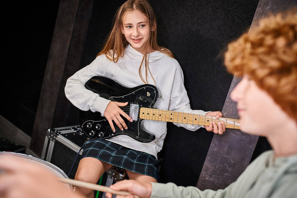 Сосредоточиться на веселой красивой девушке-подростке, держащей свою гитару и глядя на ее размытый милый барабанщик - Фото, изображение