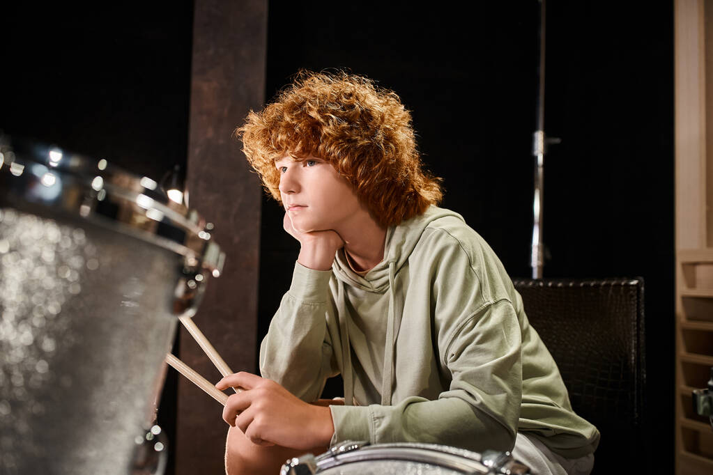 konzentrierter süßer rothaariger Teenager in lässiger Kleidung sitzt vor seinem Schlagzeug-Set - Foto, Bild
