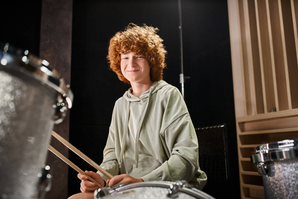 fröhlicher entzückender Teenager mit roten Haaren in lässigem Outfit vor seinem Schlagzeug, der in die Kamera lächelt - Foto, Bild