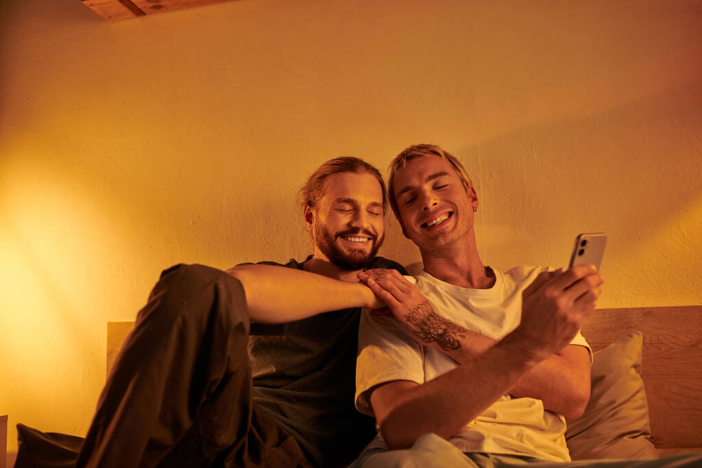 счастливый татуированный гей держит смартфон рядом с улыбающимся бородатым парнем в спальне ночью - Фото, изображение