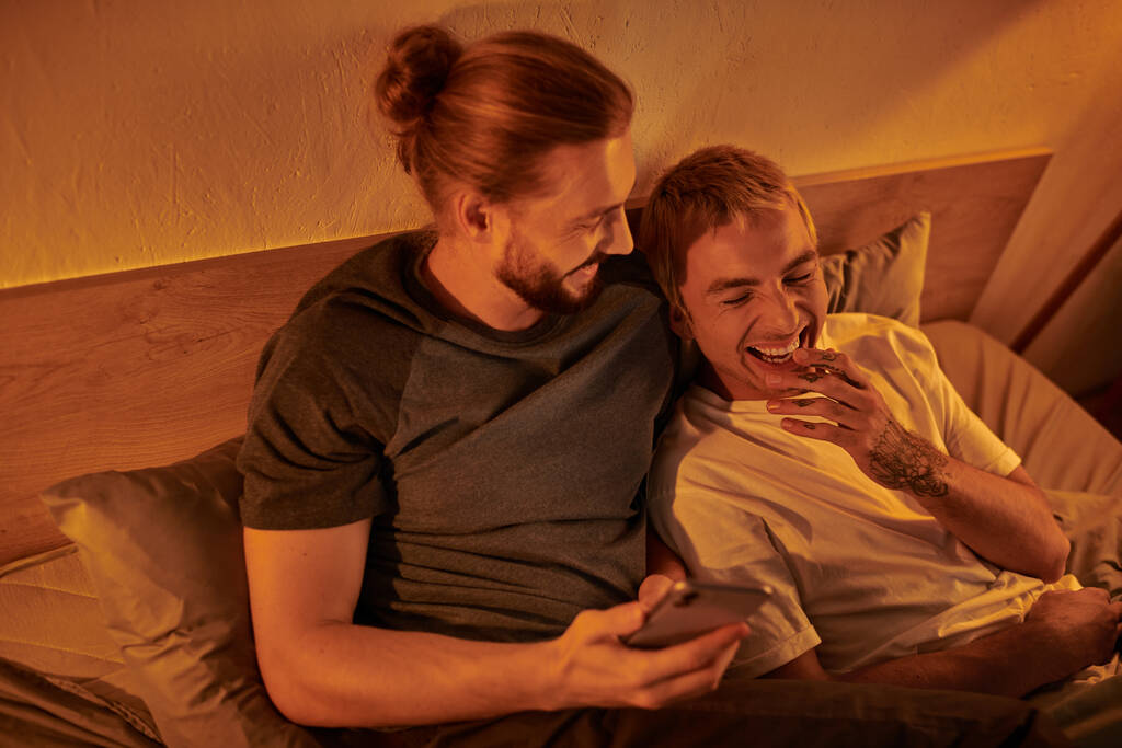 bärtiger Schwuler surft in sozialen Medien auf Smartphone neben lachendem Freund im Schlafzimmer - Foto, Bild