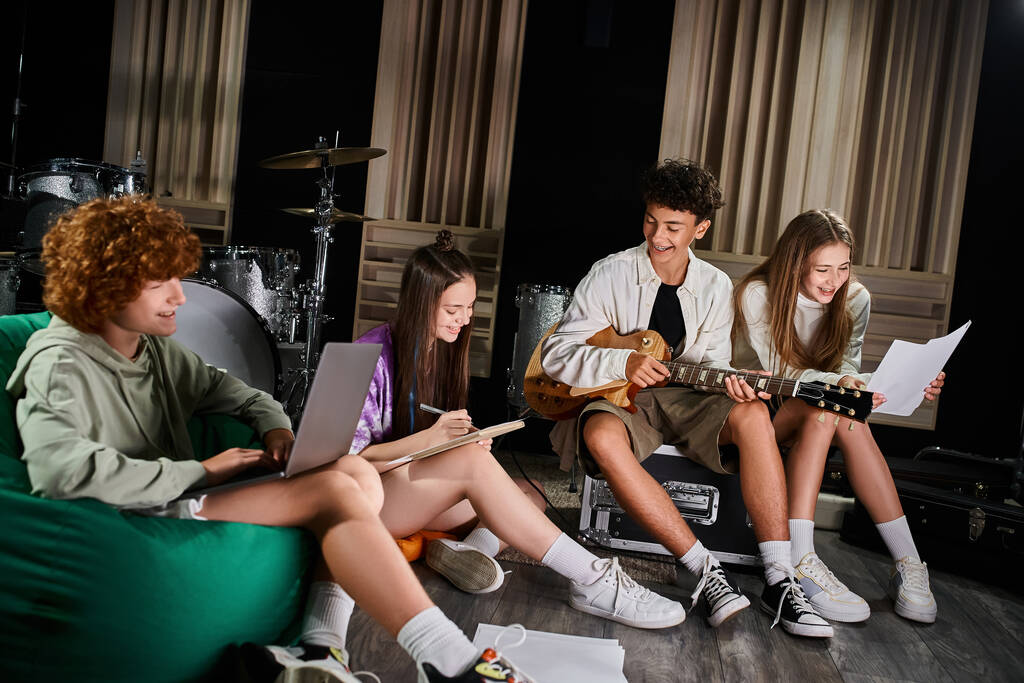 čtyři radostné teenageři členové kapely v ležérní oblečení sedí s kytarou a notebookem psaní textů - Fotografie, Obrázek