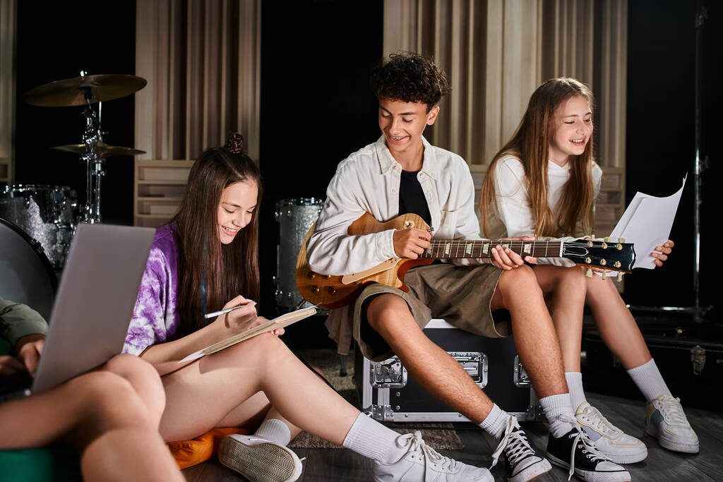 vrolijk schattig tiener band leden zitten met gitaar en laptop en het schrijven van teksten in de studio - Foto, afbeelding