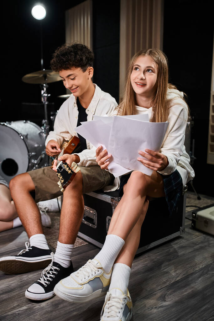 lindo adolescente tocando la guitarra cerca de su alegre amigo rubio con letras en las manos, grupo musical - Foto, imagen
