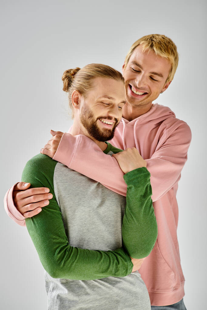 vrolijk gay paar in stijlvolle casual kleding omarmen op grijze achtergrond, harmonieuze relatie - Foto, afbeelding
