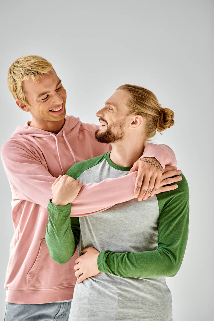 alegre pareja gay en elegante casual atuendo abrazando y mirando el uno al otro en gris telón de fondo - Foto, Imagen