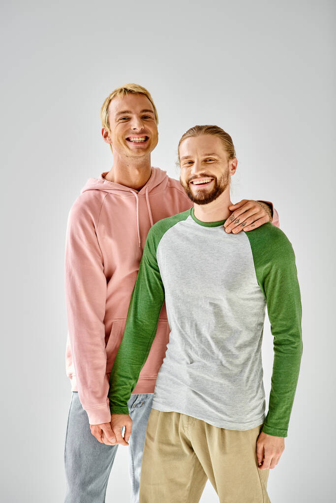 joyful gay couple in stylish casual attire standing and looking at camera on grey backdrop - Photo, Image