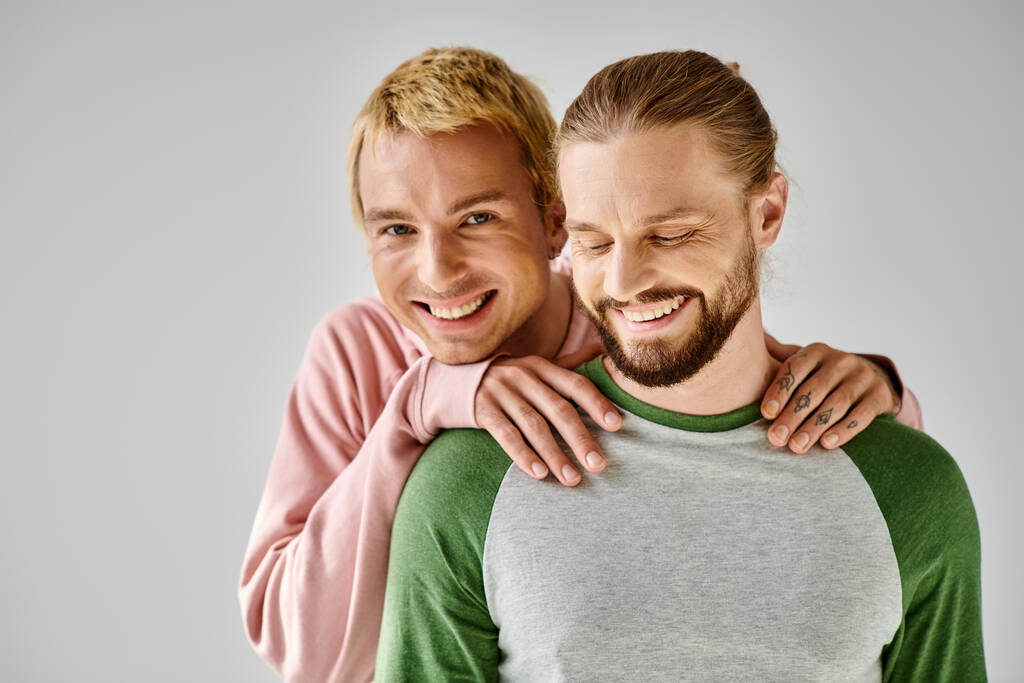 glücklich Homosexuell Mann in lässiger Kleidung umarmt Schultern von bärtigen Freund und Blick auf Kamera auf grau - Foto, Bild