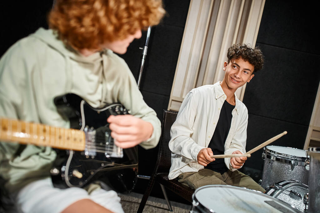 focus on jolly teenage drummer with braces smiling at his blurred red haired guitarist, musical band - Photo, Image