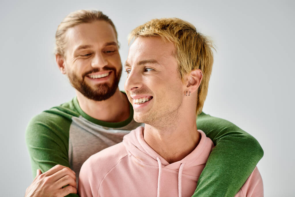 radostný vousatý gay muž objímající stylový přítel usmívající se a odvracející pohled na šedé pozadí - Fotografie, Obrázek