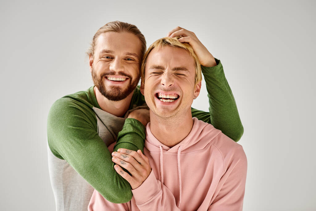 χαρούμενα γενειοφόρος γκέι άντρας αγκαλιάζει μοντέρνο φίλο, ενώ έχει τη διασκέδαση σε γκρι φόντο στο στούντιο - Φωτογραφία, εικόνα