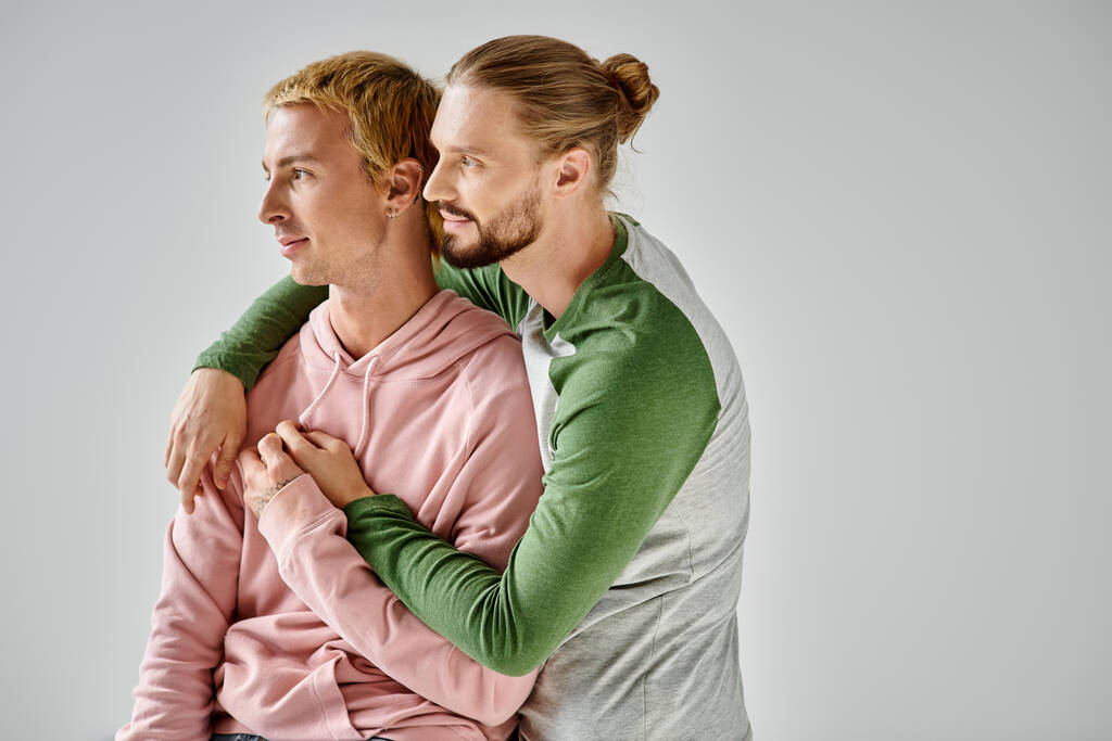ロマンチックでスタイリッシュなゲイカップルはグレーの背景に立って抱擁し,離れて見ています - 写真・画像