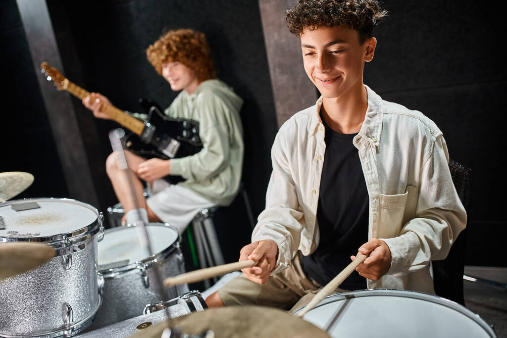 fröhlicher entzückender Teenager mit Hosenträgern, der neben seinem Freund mit Gitarre Schlagzeug spielt, Musikgruppe - Foto, Bild