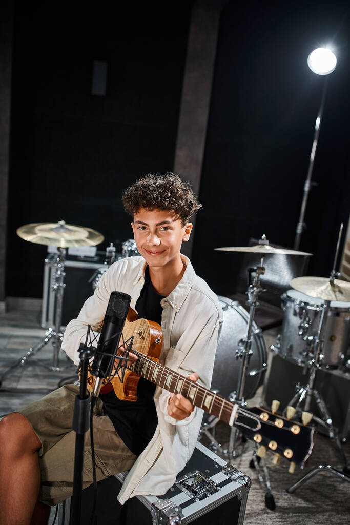 fröhlicher talentierter Teenager in lässigem Outfit, der seine Gitarre spielt und im Studio in die Kamera schaut - Foto, Bild