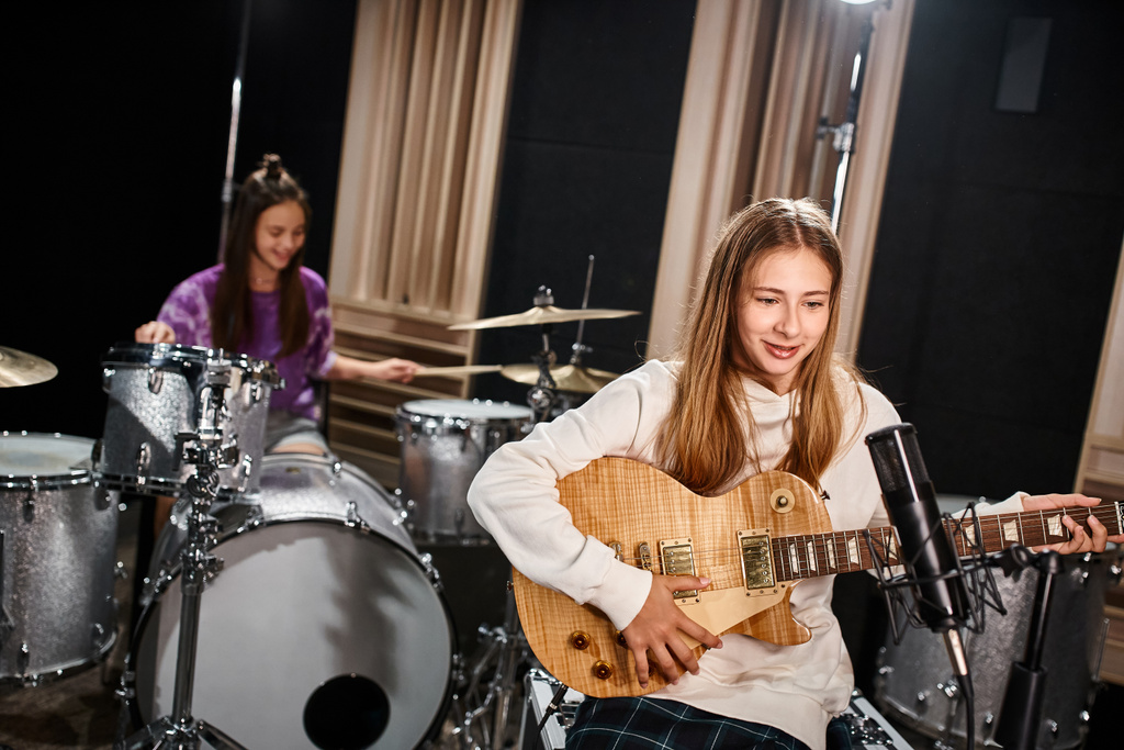 focus on pretty blonde teenage guitarist playing next to her blurred friend playing drums in studio - Photo, Image