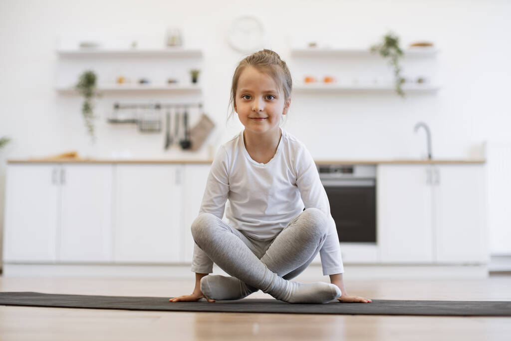 Vorderseite des netten Mädchens praktiziert Yoga, steht in Kranübung, Bakasana-Pose, trainiert auf Matte tragen Sportbekleidung, drinnen in voller Länge, in weißen Loft-Küche Hintergrund. - Foto, Bild