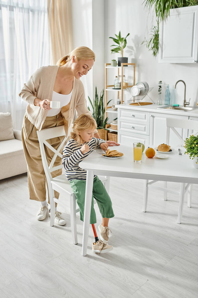 ευτυχισμένη γυναίκα στέκεται κοντά κόρη με προσθετικό πόδι έχοντας πρωινό στην κουζίνα, ένταξη - Φωτογραφία, εικόνα