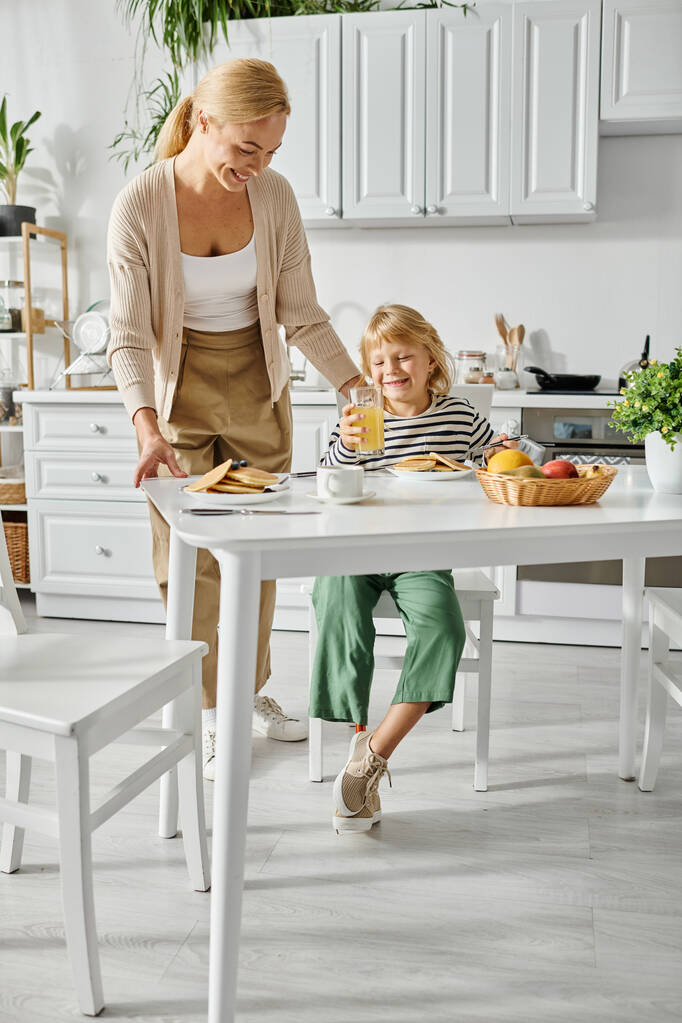 キッチンで朝食中に義足で娘にパンケーキを提供する幸せな女性, 含める - 写真・画像
