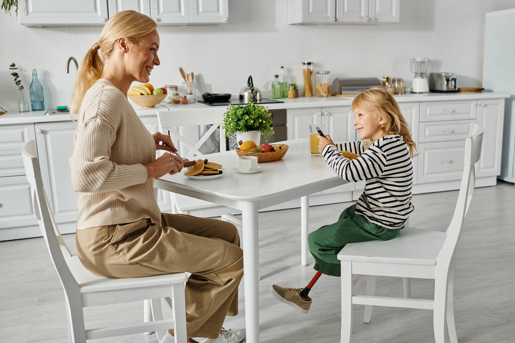 feliz madre desayunando con su hijita con prótesis en la cocina, inclusión - Foto, Imagen