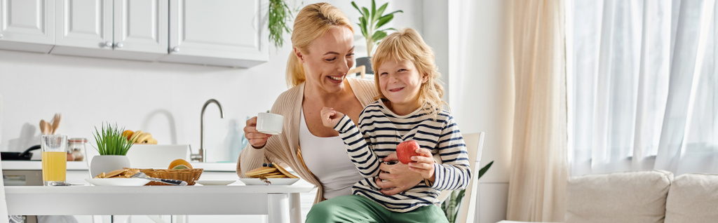 fröhliches Mädchen mit Apfel in der Hand und auf dem Schoß einer glücklichen Mutter beim Frühstück in der Küche, Transparent - Foto, Bild
