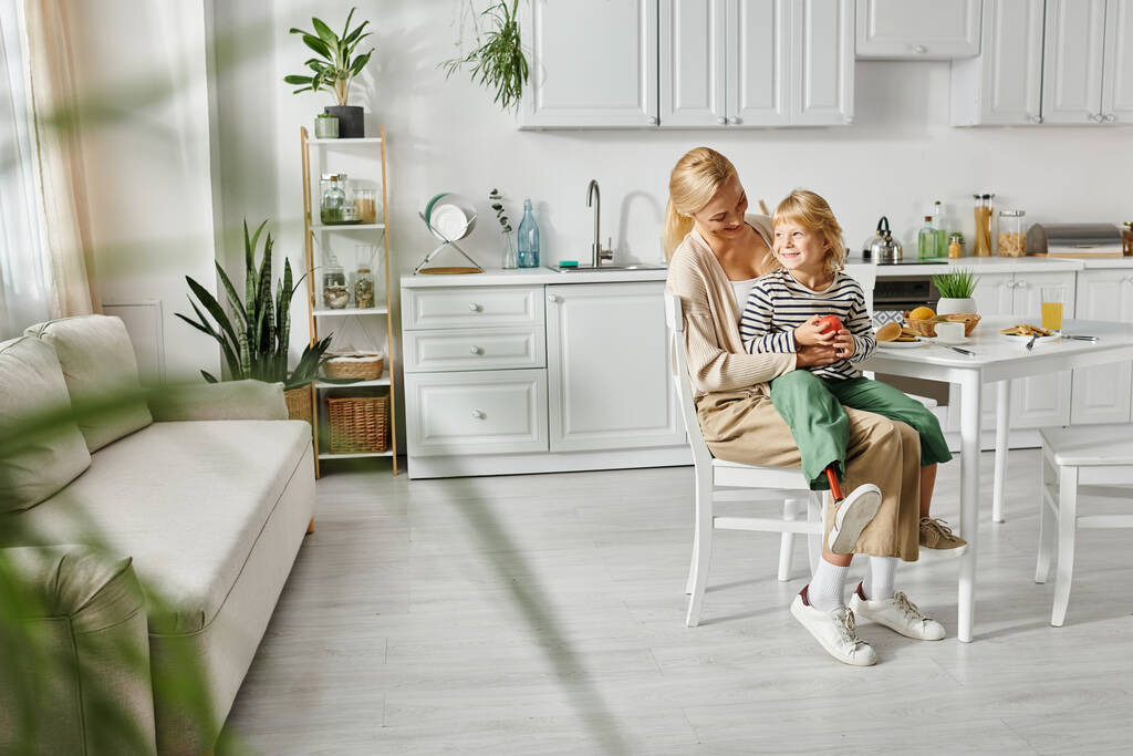 Kleines Mädchen mit Beinprothese auf dem Schoß einer glücklichen blonden Mutter beim Frühstück in der Küche - Foto, Bild