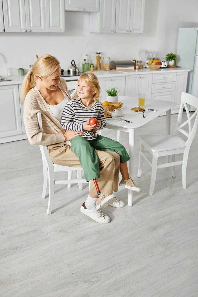 niña en edad preescolar con la pierna protésica sentado en vueltas de madre feliz durante el desayuno en la cocina - Foto, Imagen