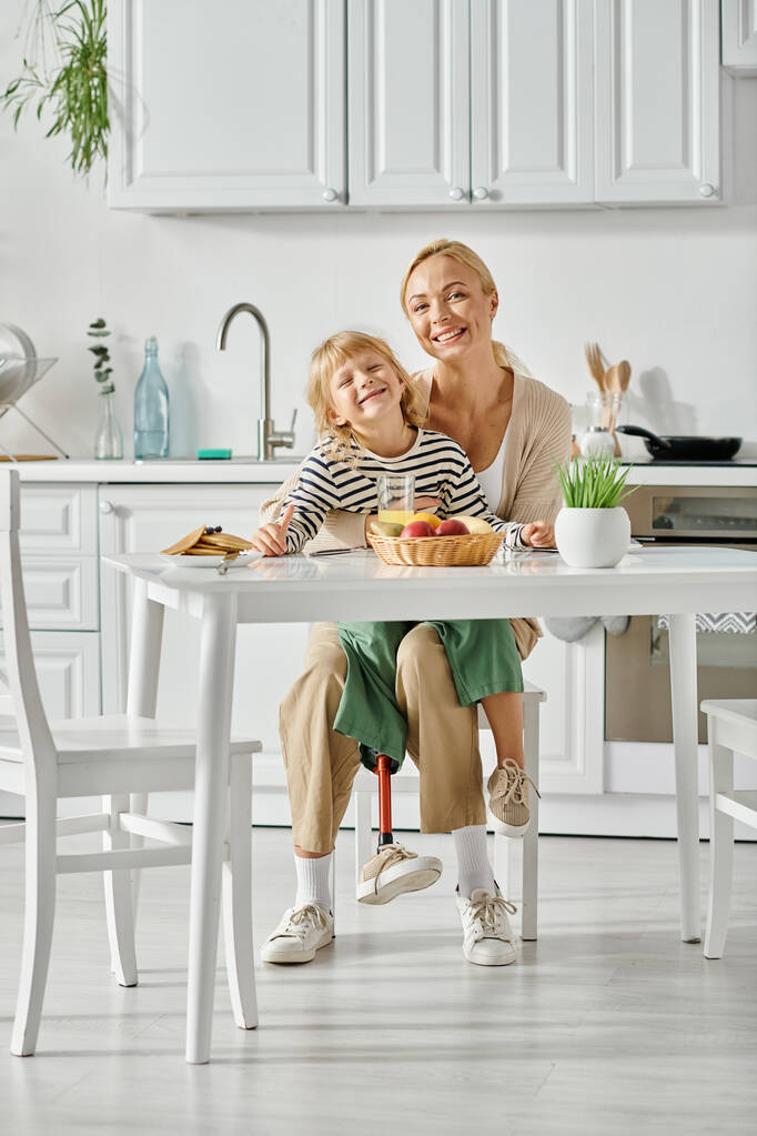 menina alegre com a perna protética sentado em voltas de mãe feliz durante o café da manhã na cozinha - Foto, Imagem