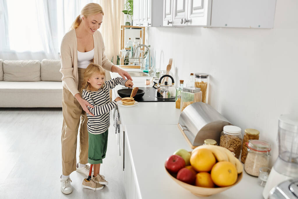 mãe feliz de pé perto pequena filha com prótese perna fritando panquecas na cozinha moderna - Foto, Imagem