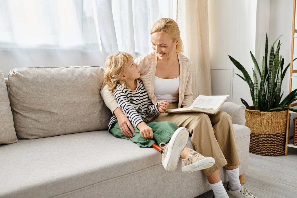 feliz madre leyendo el libro a su hija con la pierna protésica y sentados juntos en la sala de estar - Foto, Imagen