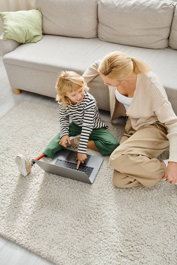 gelukkig kind met prothese been wijzen naar laptop en zitten op tapijt met moeder in de woonkamer - Foto, afbeelding