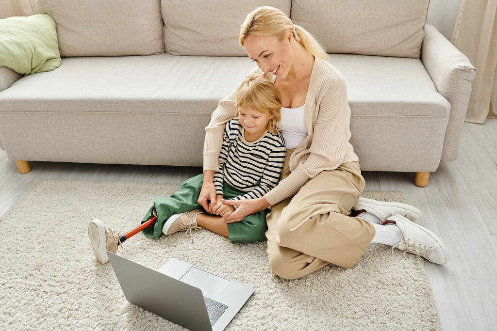 szczęśliwe dziecko z protezą nogi oglądając film na laptopie siedząc na dywanie z matką - Zdjęcie, obraz