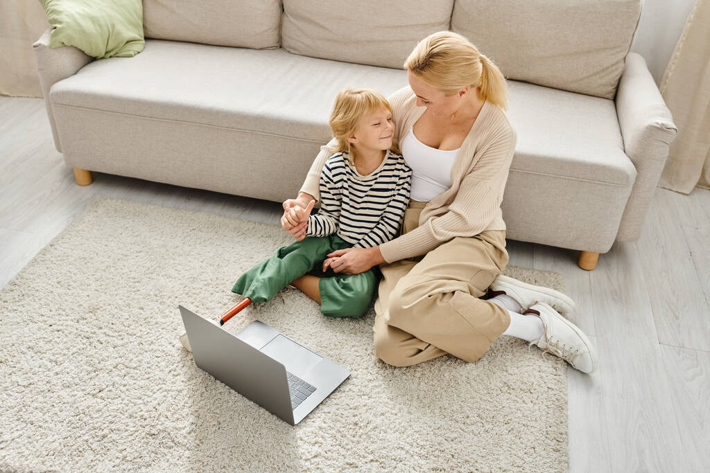 счастливая девушка с протезной ногой смотрит фильм на ноутбуке и сидит на ковре вместе с мамой - Фото, изображение
