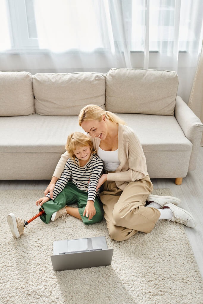 счастливый ребенок с протезной ногой смотреть фильм на ноутбуке и сидеть на ковре вместе с матерью - Фото, изображение