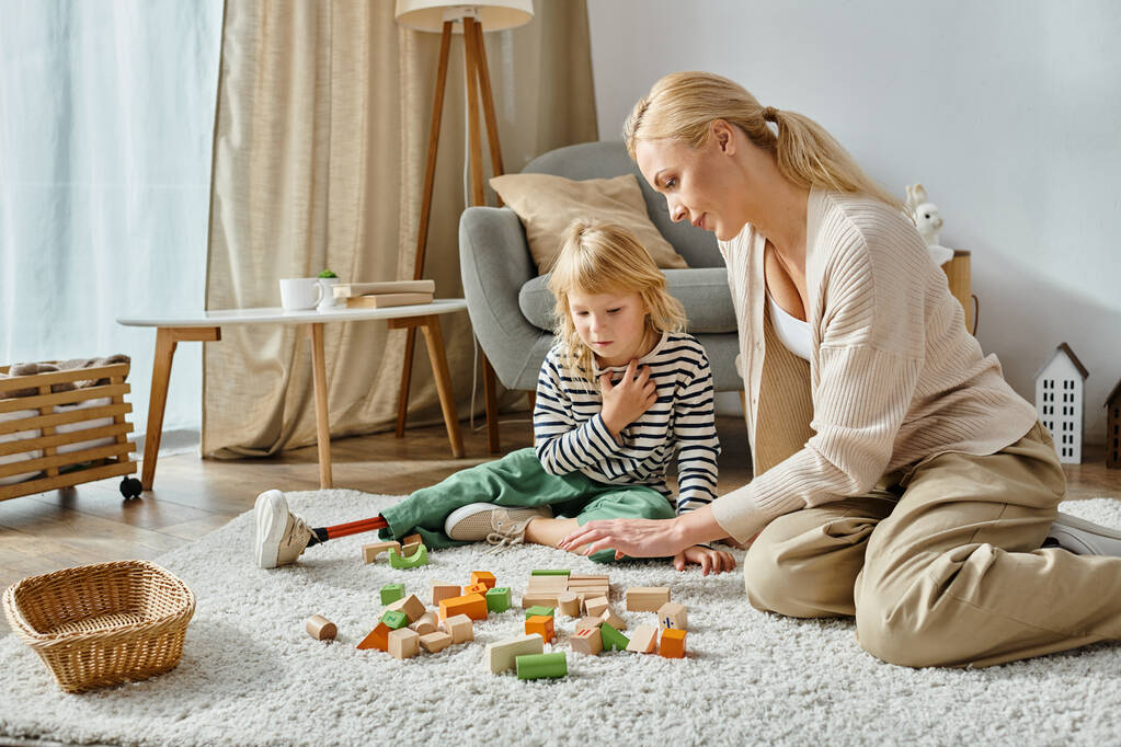 fille handicapée avec prothèse jambe assise sur le tapis et en regardant des jouets en bois près de la mère - Photo, image