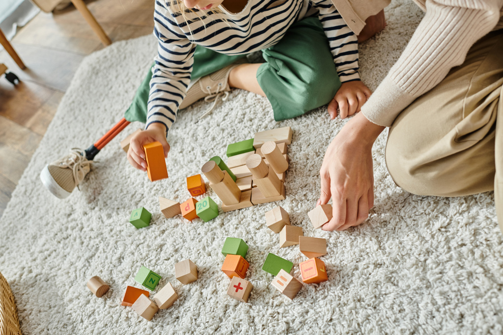 Mädchen mit Beinprothese sitzt auf Teppich und spielt mit Holzspielzeug in der Nähe der Mutter - Foto, Bild