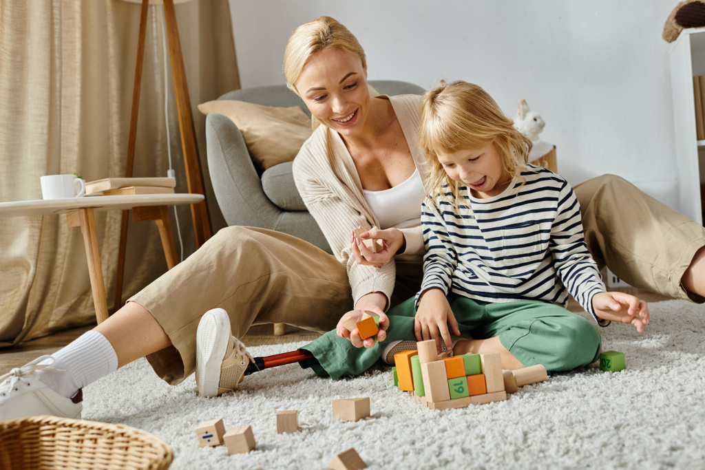 niña preescolar con la pierna protésica sentado en la alfombra y jugando con bloques de madera cerca de la madre - Foto, imagen