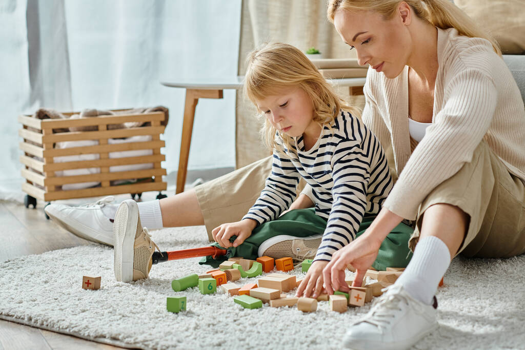 fille blonde avec prothèse jambe assise sur le tapis et jouer avec des blocs de bois éco près de la mère - Photo, image