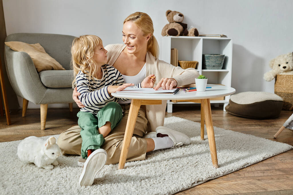 happy blonde woman looking at her daughter with prosthetic leg drawing on paper with colorful pencil - Photo, Image