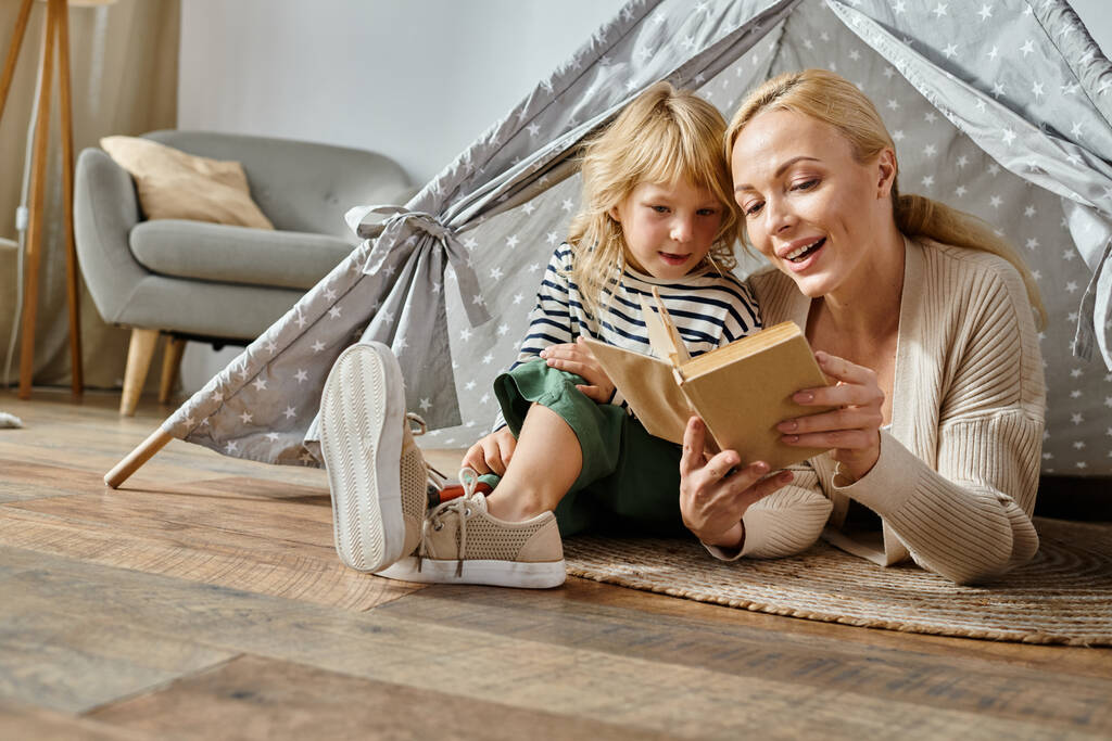 carino ragazza con protesi gamba e bionda madre lettura libro e seduto in tenda da gioco a casa - Foto, immagini