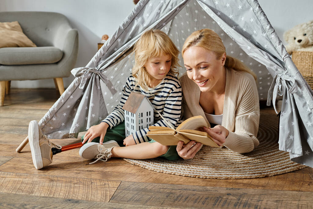 Nettes Mädchen mit Beinprothese und blonder Mutter, die Buch liest und zu Hause im Spielzelt sitzt - Foto, Bild