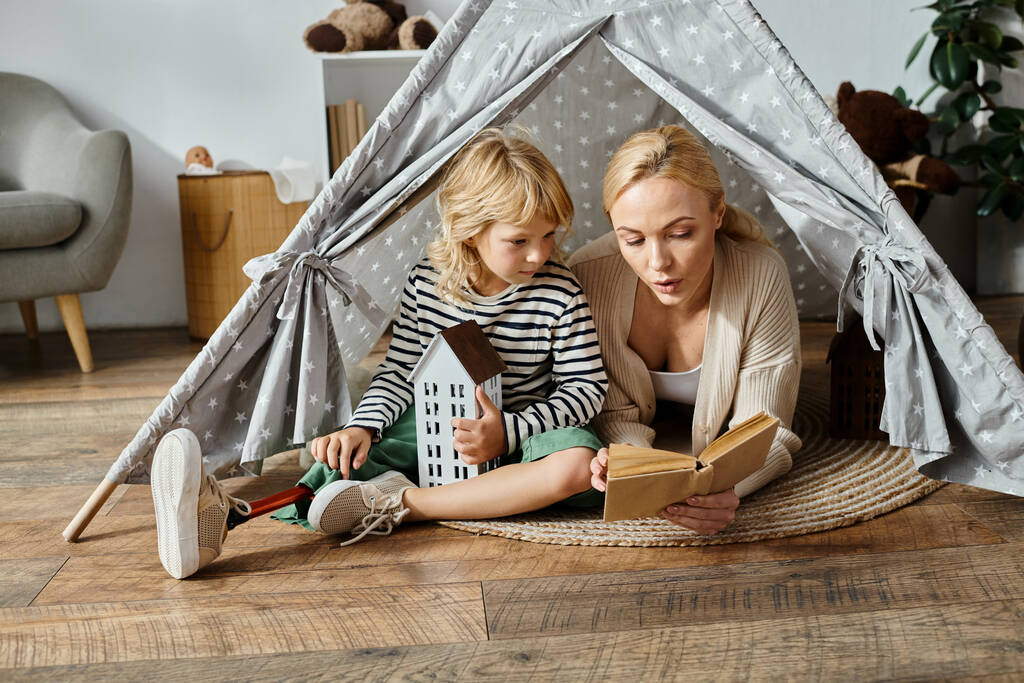 słodkie dziewczyna z protezy nogi i blondynka matka czytania książki siedząc w namiocie zabaw w domu - Zdjęcie, obraz